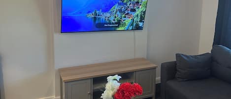 A living room with a flat-screen TV mounted on the wall, a wooden coffee table with a vase of flowers, a remote control, and a grey sofa.