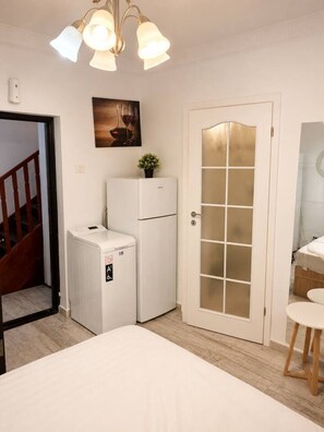 Un dormitorio con cama, refrigerador pequeño, puerta con paneles de vidrio esmerilado, lámpara de techo y un cuadro en la pared.