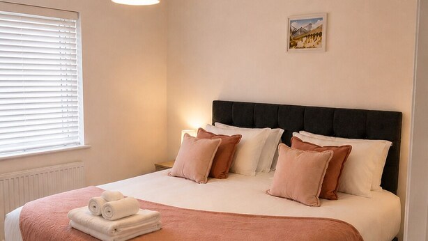 Una camera da letto ordinata con un letto, cuscini, un comodino, una lampada e un termosifone.