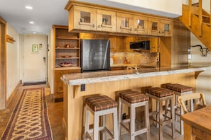 A kitchen with wooden cabinets, a stainless steel refrigerator, a microwave, and a marble countertop. There are three bar stools with plaid cushions. - Columbine 2308 (Crested Butte)