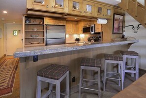 A kitchen with wooden cabinets, a stainless steel refrigerator, a microwave, and a bar area with stools. - Columbine 2308 (Crested Butte)
