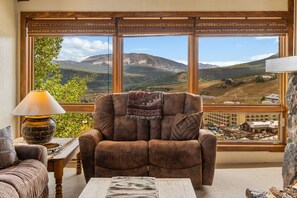 A room with a large window offering a mountain view, a brown leather armchair with a patterned throw, a wooden coffee table, and a lamp. - Columbine 2308 (Crested Butte)