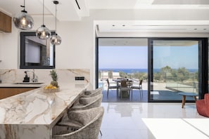 A modern kitchen with a marble countertop, a dining area with a view of the ocean, and a large sliding glass door. - MT7 Panoramic Sea View Villa, Villa (Limenas Chersonisou)