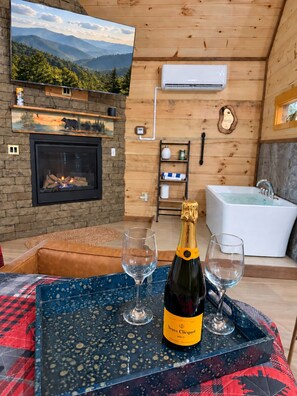 Ein gemütliches Zimmer mit Kamin, einem großen Fenster mit schöner Aussicht, einer Badewanne und einer Champagnerflasche auf einem Tablett.