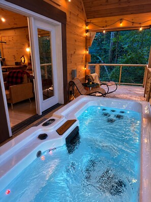 A hot tub with a wooden deck, a glass door leading to a room, and string lights. - Romantic Cabin • Fireside Jacuzzi + Hot Tub (Sevierville)