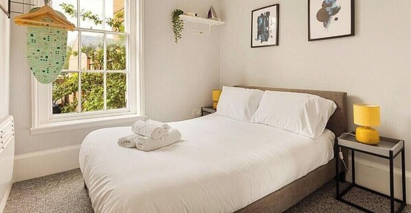 A neatly made bed with white linens, a bedside table with a lamp, and a window with a view of greenery outside. - Relaxing 3BR | Near Local Stores | Relocation |ST (Oxfordshire)