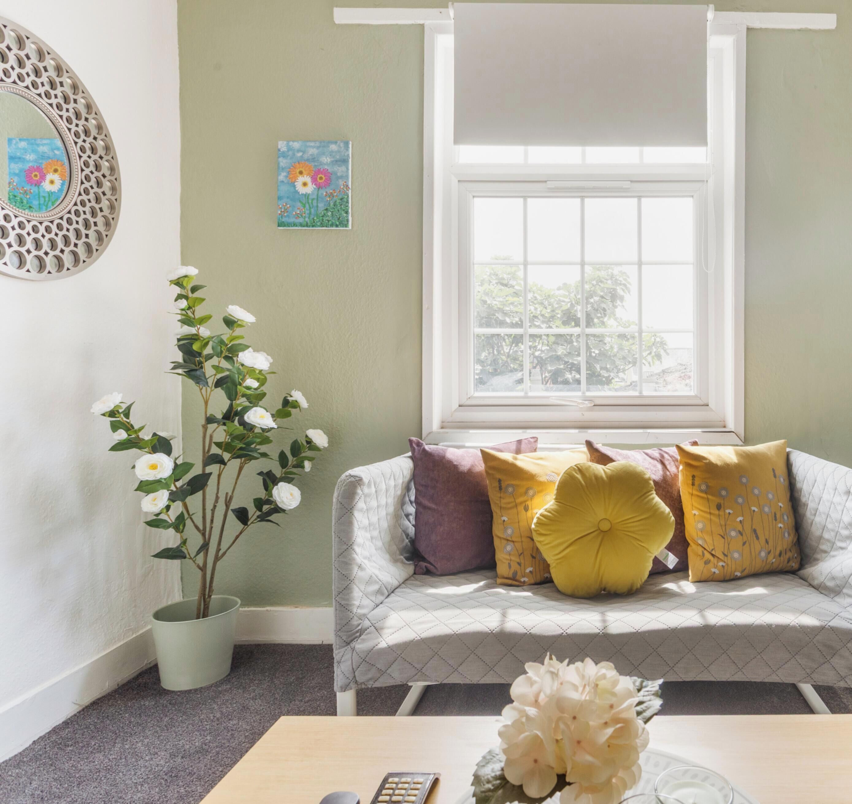 A room with a window, a sofa with cushions, a potted plant, and a table with a vase.