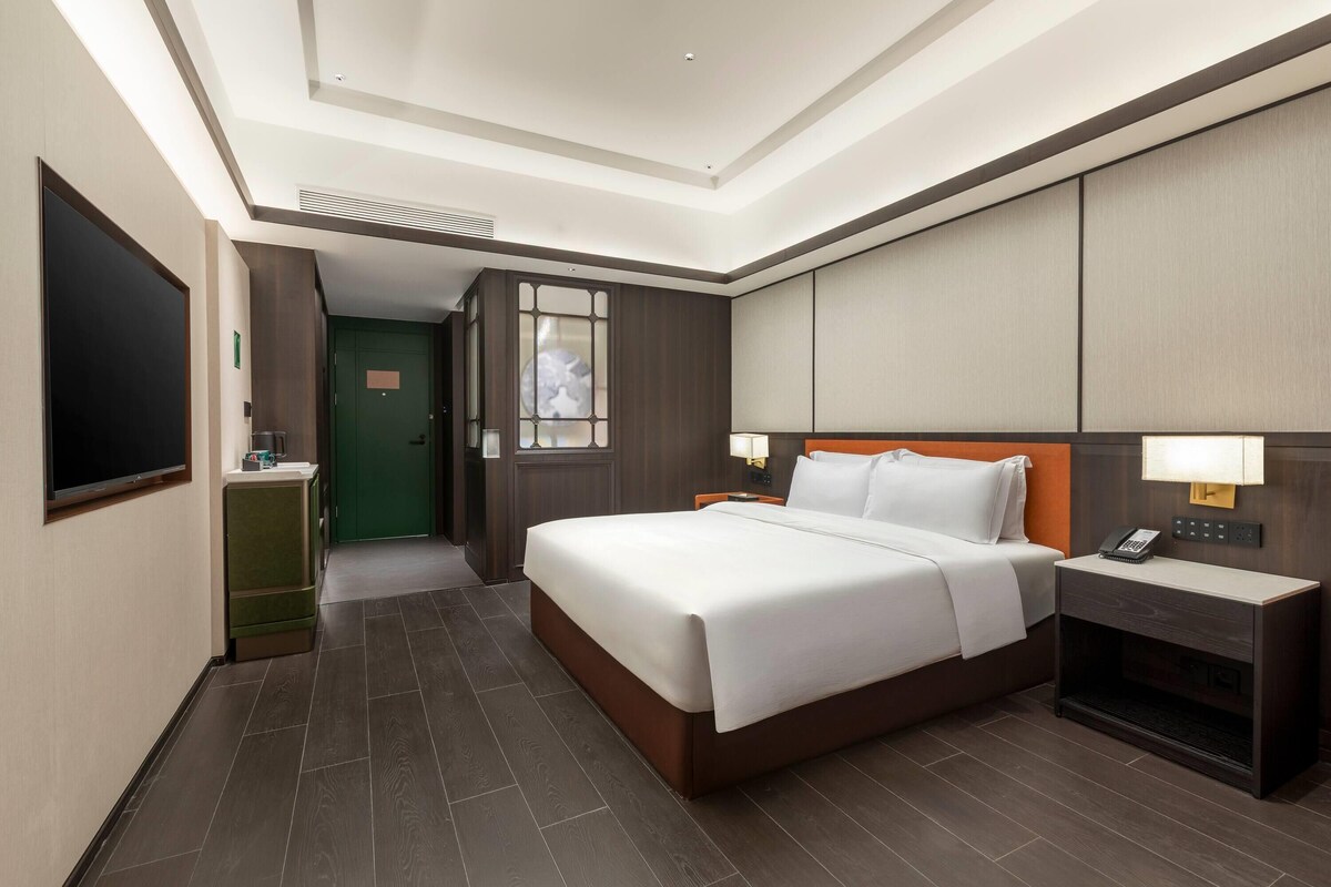 A hotel room with a large bed, bedside tables, a flat-screen TV, a green door, and a view of the hallway.