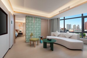 Habitación de hotel moderna con sofá, una mesita, una silla, televisor y vistas a la ciudad a través de grandes ventanales.