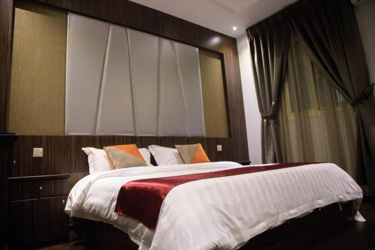 Ein Hotelzimmer mit einem großen Bett, einem hölzernen Kopfteil und Blick auf die Stadt durch transparente Vorhänge.