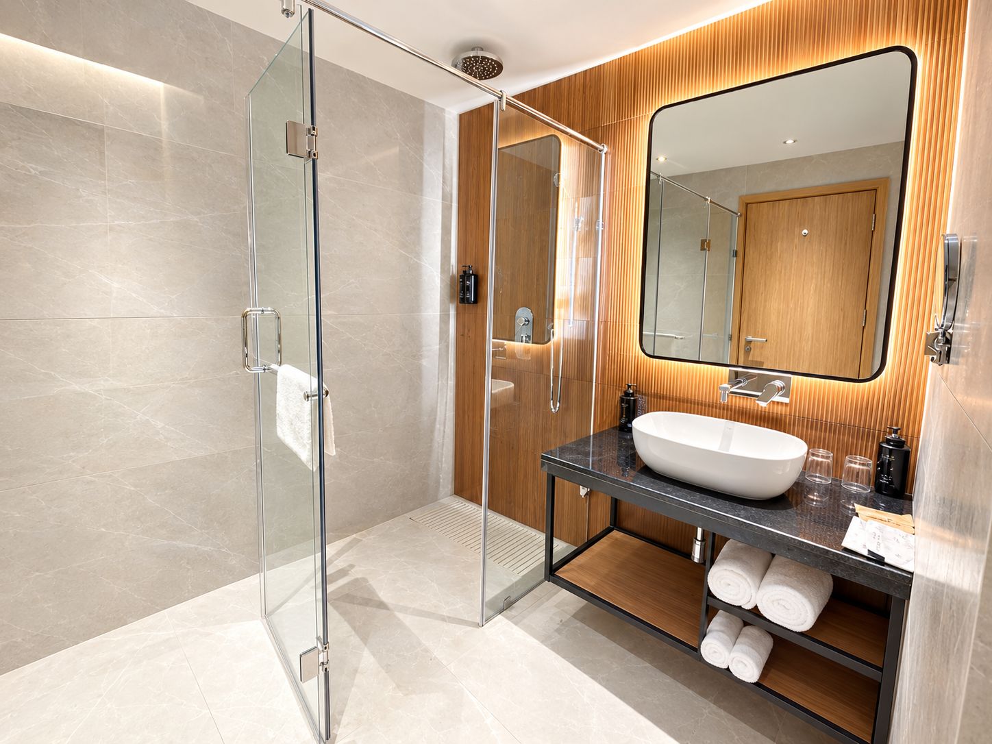 A modern bathroom with a glass shower enclosure, a wooden vanity with a large mirror, and neatly rolled towels.