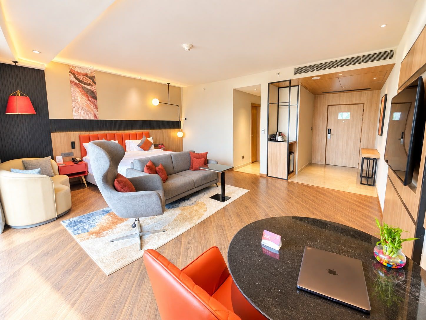 A modern hotel room with a sofa, armchair, and a dining area with a laptop and a vase.