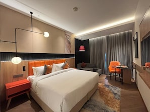 A hotel room with a large bed, bedside tables, a desk, a chair, and a window with curtains. - Ramada Encore By Wyndham Ayodhya Takpura (Faizabad)