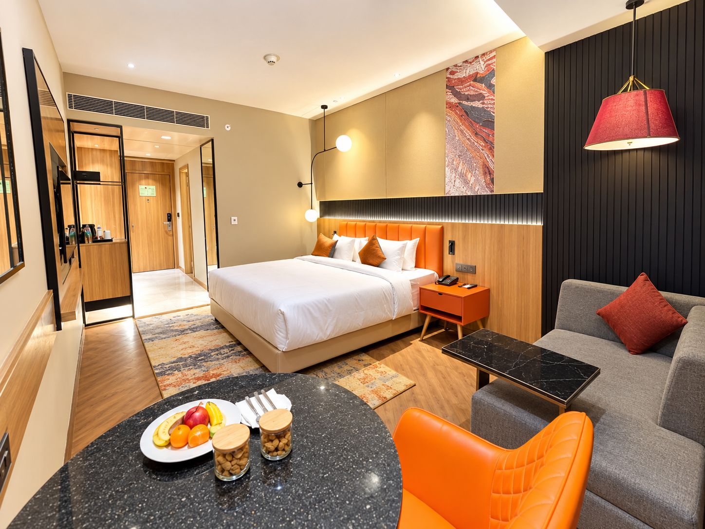 A hotel room with a bed, a sofa, a dining table with fruit and nuts, and a red lamp.