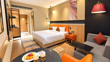 A hotel room with a bed, a sofa, a dining table with fruit and nuts, and a red lamp.