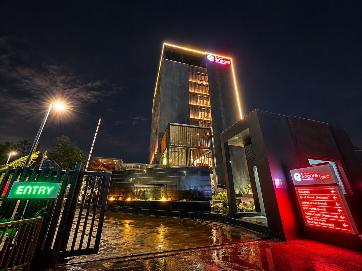 Entrada de hotel moderna con un letrero iluminado, un mostrador de recepción y el suelo mojado reflejando luces.