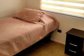 Een enkel bed met een geruit sprei, een nachtkastje met lades en een raam met jaloezieën.