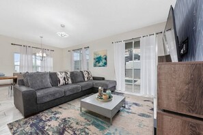 A modern living room with a grey sofa, a coffee table, and a large window with white curtains. - 5BR Storey Lake Home Near Disney With Pool (Kissimmee)