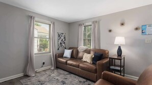Een woonkamer met een bruine leren bank, een kussen met een patroon, een klein bijzettafeltje met een lamp, en een bord met tekst aan de muur.