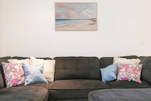 A dark-colored sofa with multiple pillows, a painting of a beach scene, and a plain wall. - Tropical Breeze Poolside Stay near the Waterfront (Parap)