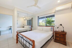 A bedroom with a bed, bedside table, mirror, window with view, ceiling fan, and air conditioning unit. - Tropical Breeze Poolside Stay near the Waterfront (Parap)
