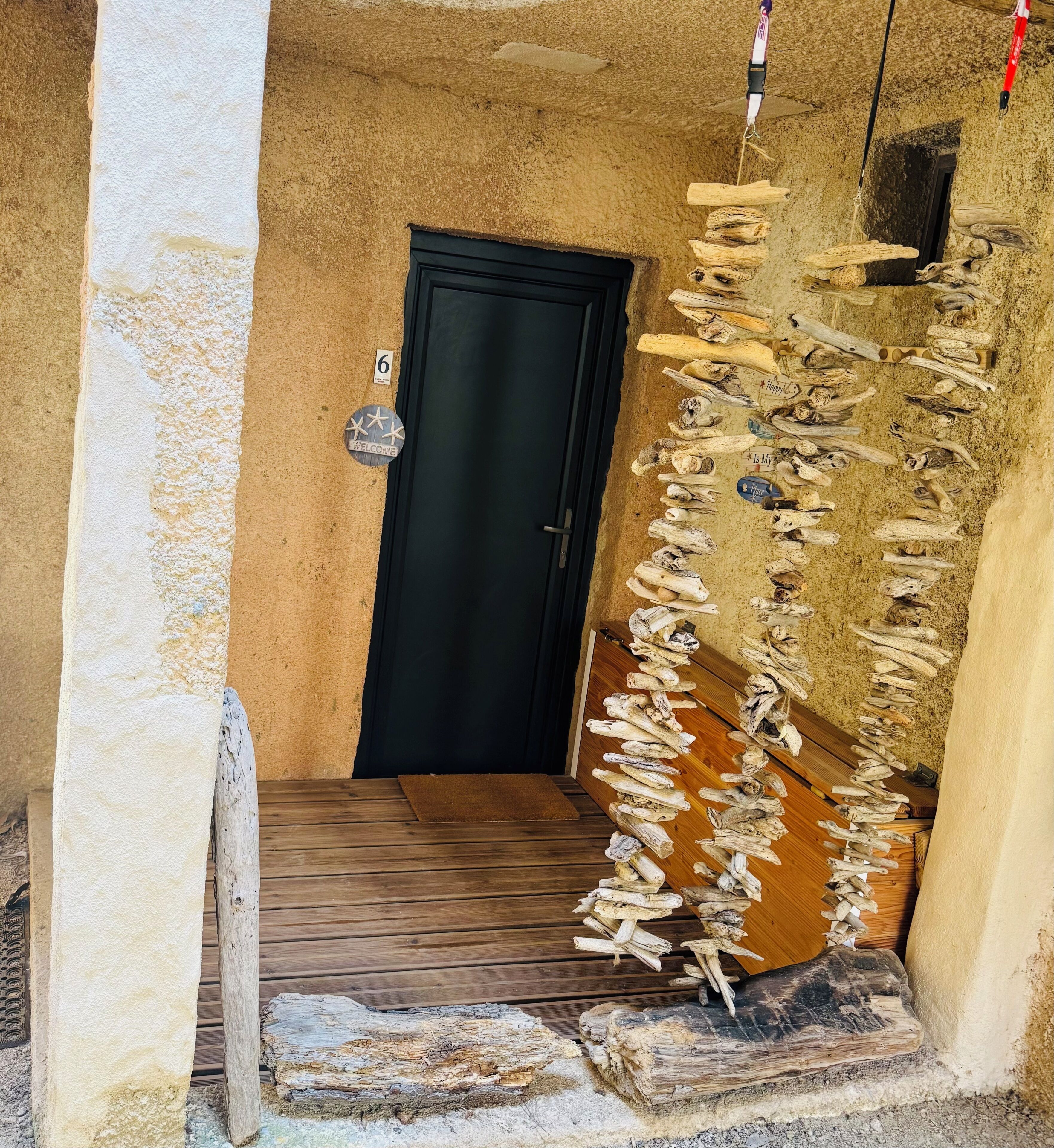 Un couloir étroit avec une porte en bois, une pancarte de bienvenue et un mur décoré de morceaux de bois flotté.