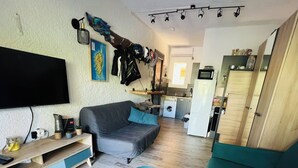 A living room with a grey sofa, a teal sofa, and two stools, a flat-screen TV, a kitchen area with a washing machine, and a ceiling-mounted light fixture. - Perfect for couples—just a 3-minute walk from Liscia Beach. Pets allowed. (Calcatoggio)