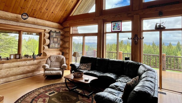 A log cabin interior with a leather sofa, a chair, a table, and large windows offering a forest view.