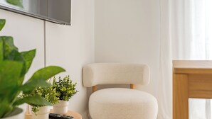 Ein modernes Wohnzimmer mit einem weißen Sessel, einem Holztisch mit Fernbedienung und Topfpflanzen sowie einem an der Wand montierten Fernseher.