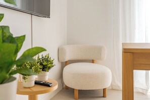 Ein modernes Wohnzimmer mit einem weißen Sessel, einem Holztisch mit Fernbedienung und Topfpflanzen sowie einem an der Wand montierten Fernseher.