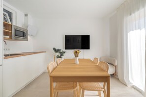 A modern kitchen with white cabinets, a microwave, and a built-in oven. There is a dining table with six wicker chairs and a vase with flowers. A flat-screen TV is mounted on the wall above the table. - Sunny Guargacho6 Retreat—South Tenerife (Guargacho)
