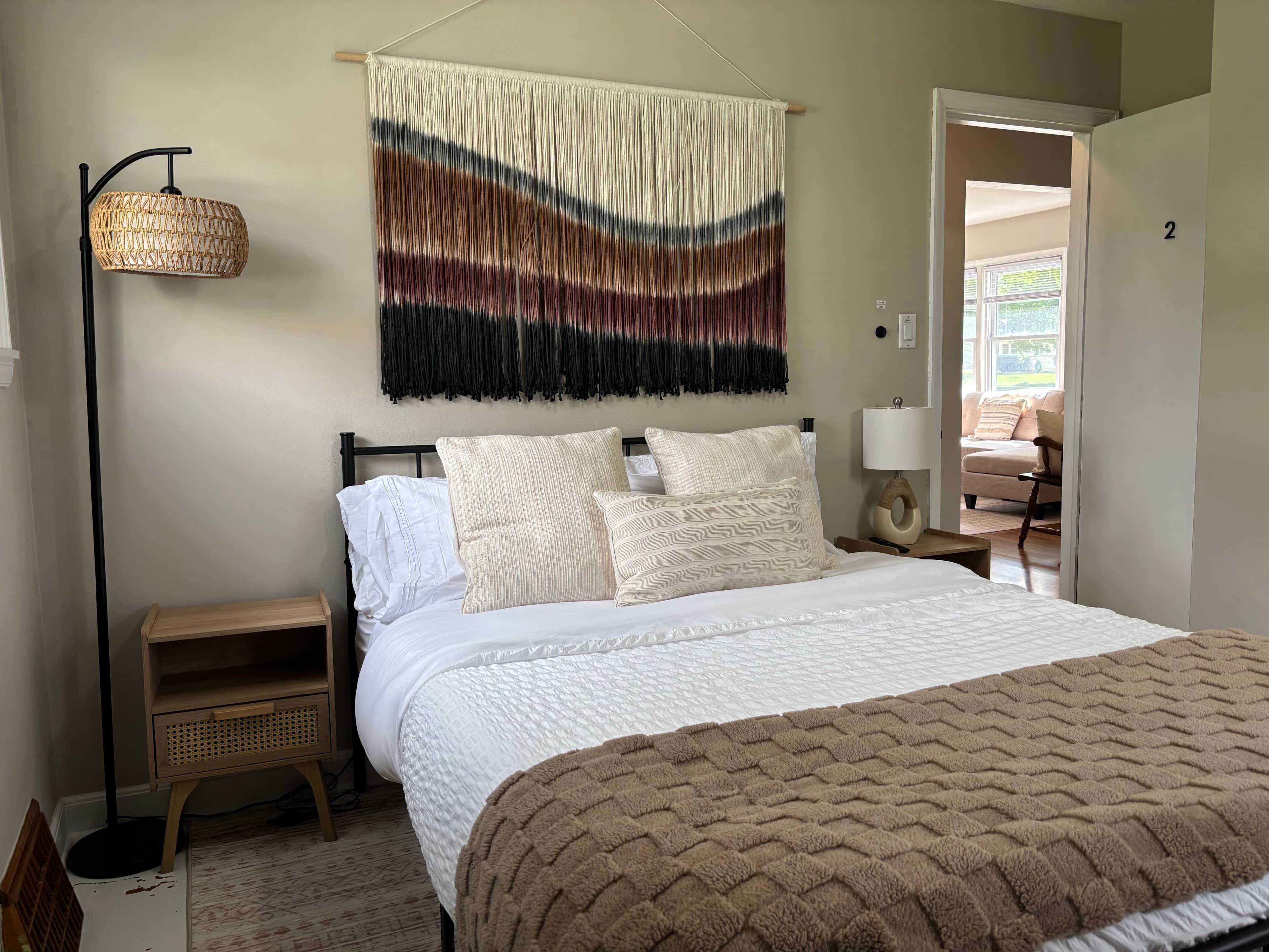 Une chambre à coucher avec un lit, des tables de chevet, une décoration murale en macramé et une vue sur l’extérieur.