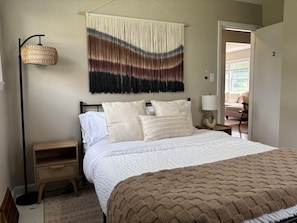 A bedroom with a bed, bedside tables, a hanging macramé wall hanging, and a view of the outside.