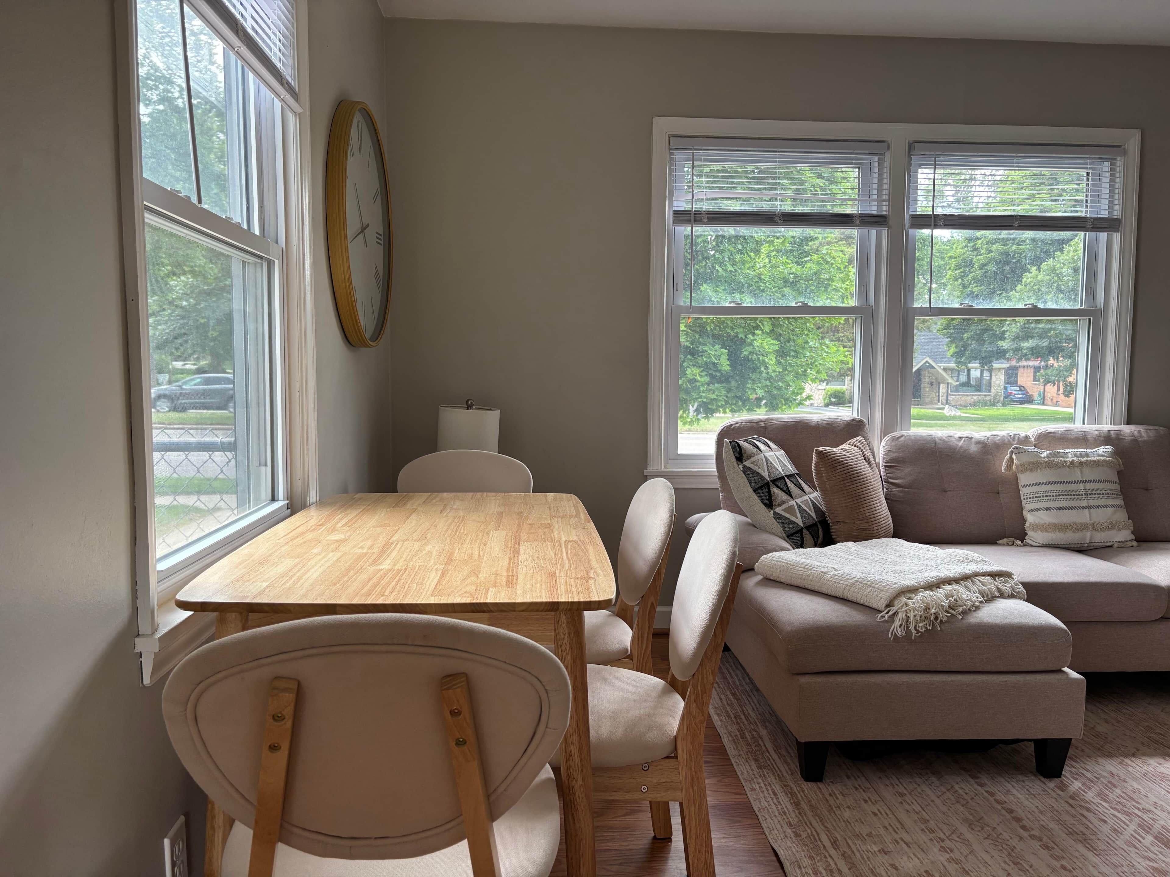 Un salon avec une table en bois, un canapé et un fauteuil. Il y a deux fenêtres avec des stores, une horloge au mur et une lampe sur la table.