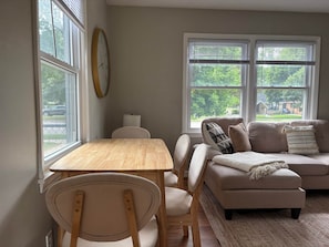 A living room with a wooden table, a sofa, and a chair. There are two windows with blinds, a clock on the wall, and a lamp on the table.