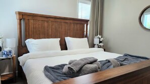 Ein ordentlich bezogenes Bett mit Holz-Kopfstück, ein Nachttisch mit Lampe und Taschentuchbox sowie ein Fenster mit Vorhängen.