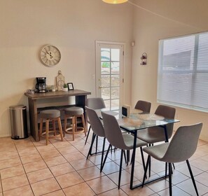 Een eetruimte met een glazen tafel, grijze stoelen, een koffieapparaat en een klok aan de muur.