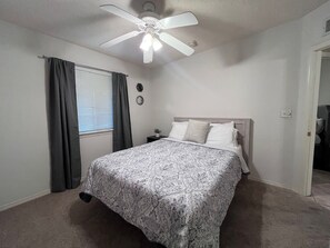 Une chambre à coucher comprenant un lit, un ventilateur de plafond, une fenêtre avec des rideaux, une table de chevet et une horloge murale.