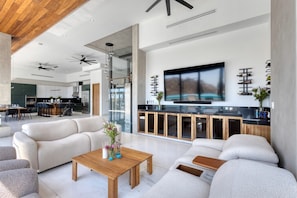 A modern living room with a large TV, sofas, a coffee table, and a kitchen in the background. - New Modern Estate 20Bd, Gym, Sauna, Massage, Games (Cabo San Lucas)