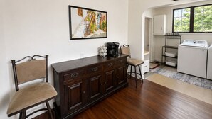 A room with a dark wooden cabinet, two chairs, a coffee maker, and a painting on the wall.