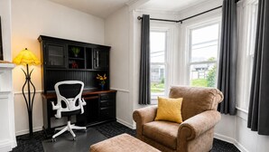 A room with a desk, chair, ottoman, and a window with curtains.