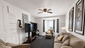 A living room with a TV, two sofas, a chair, a ceiling fan, and framed pictures on the walls.