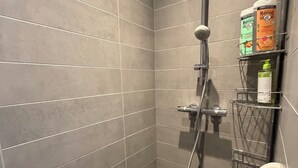 Een douche met grijze tegels, een handdouche, een handdoucheslang, een metalen plank met toiletartikelen, en een veiligheidsbeugel.