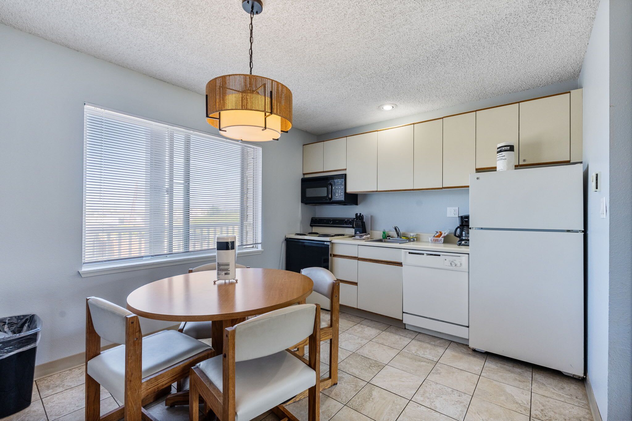 A compact kitchen with a dining table, chairs, a microwave, a refrigerator, and a hanging light fixture.