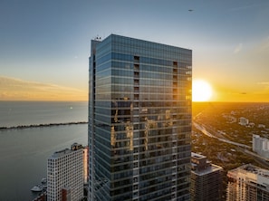 En høj bygning med glasfacade, beliggende nær et vandareal med solnedgang i baggrunden.