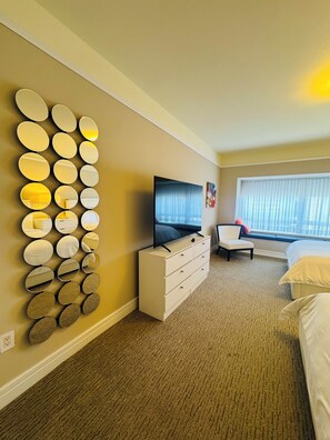 Een hotelkamer met een spiegel aan de muur, een flatscreen televisie, een witte dressoir, een stoel, een bed en een groot raam met gordijnen.