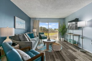 Ein Wohnzimmer mit Sofa, Couchtisch, Lampe, Fernseher und Blick durch eine Schiebetür.
