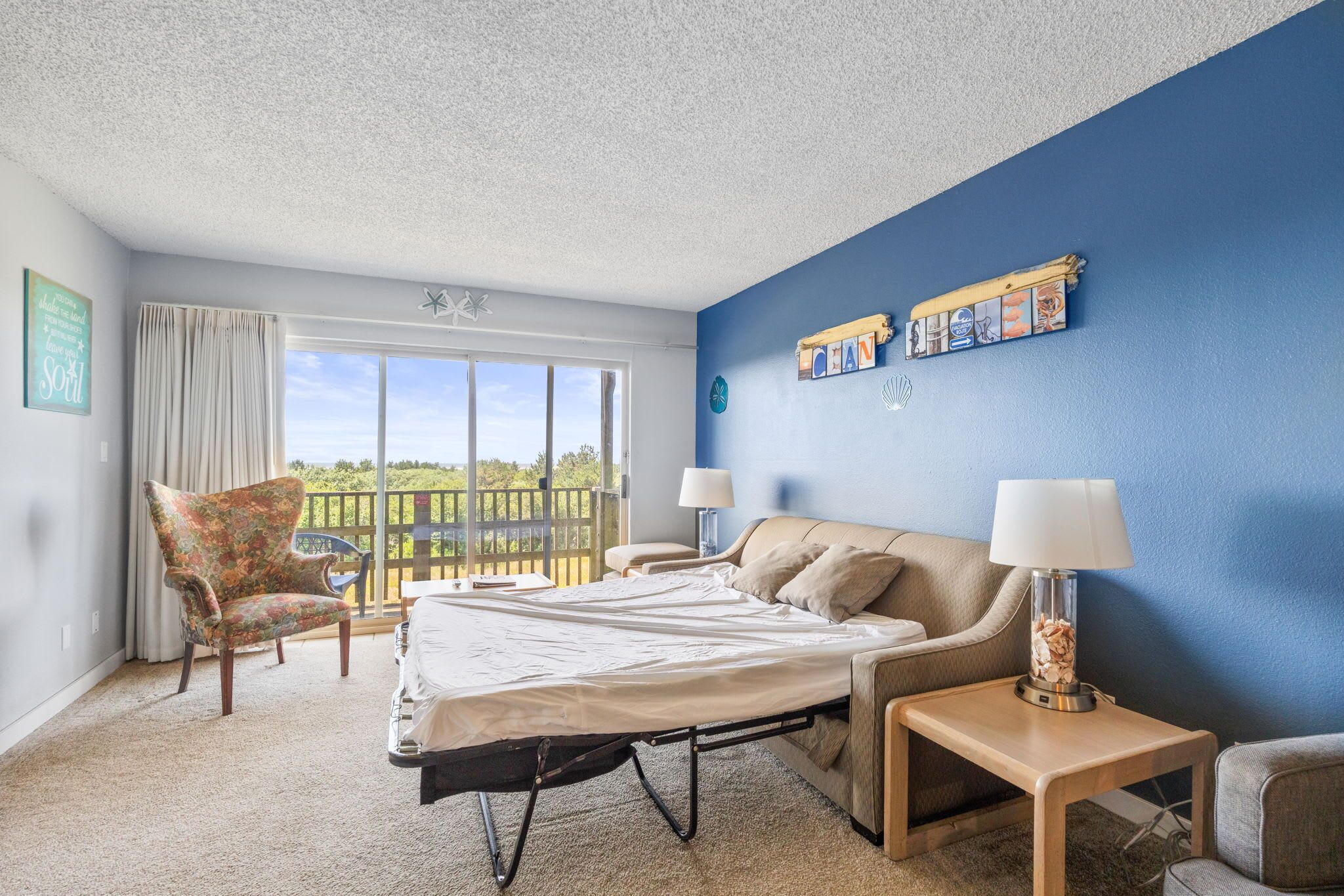 A hotel room with a bed, a chair, a nightstand, a lamp, a view of the outdoors, and a blue wall.