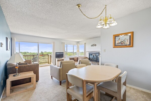 A spacious living room with a large dining table, comfortable seating, a television, and a balcony with a view.