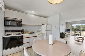 A compact kitchen with a microwave, stove, and refrigerator, a round dining table with chairs, and a balcony with a TV and seating area. - Pet-Friendly One-Bedroom Condo - Pool & Hot Tub! (Ocean Shores)